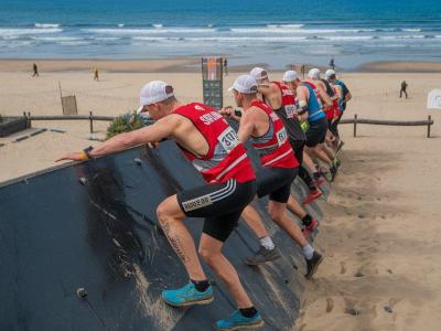 Atleti scalano un muro durante una gara sulla spiaggia.