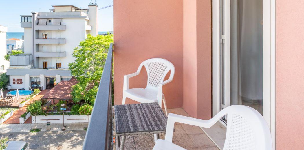 Balcone con sedie bianche, tavolino, vista su edificio e mare in lontananza.