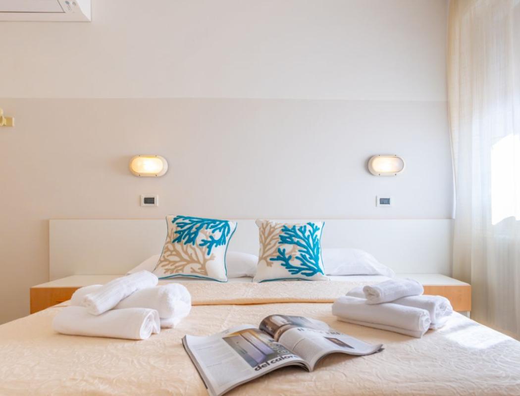 Camera luminosa con letto matrimoniale, cuscini decorativi e asciugamani piegati su copriletto.