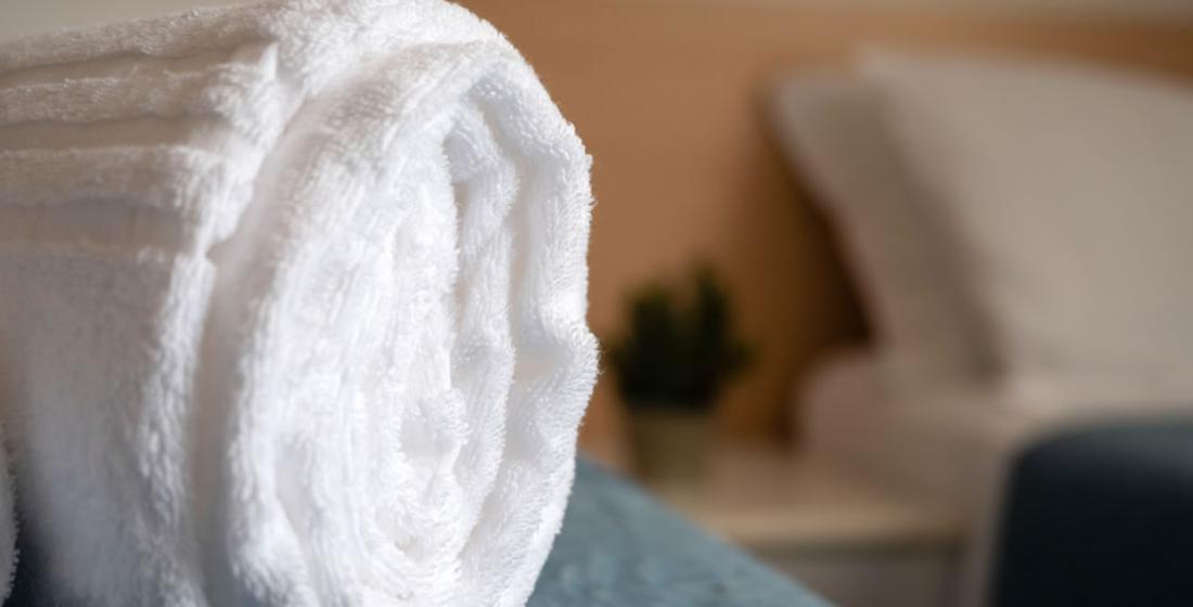 Asciugamano bianco arrotolato su un letto, con sfondo sfocato di una camera.