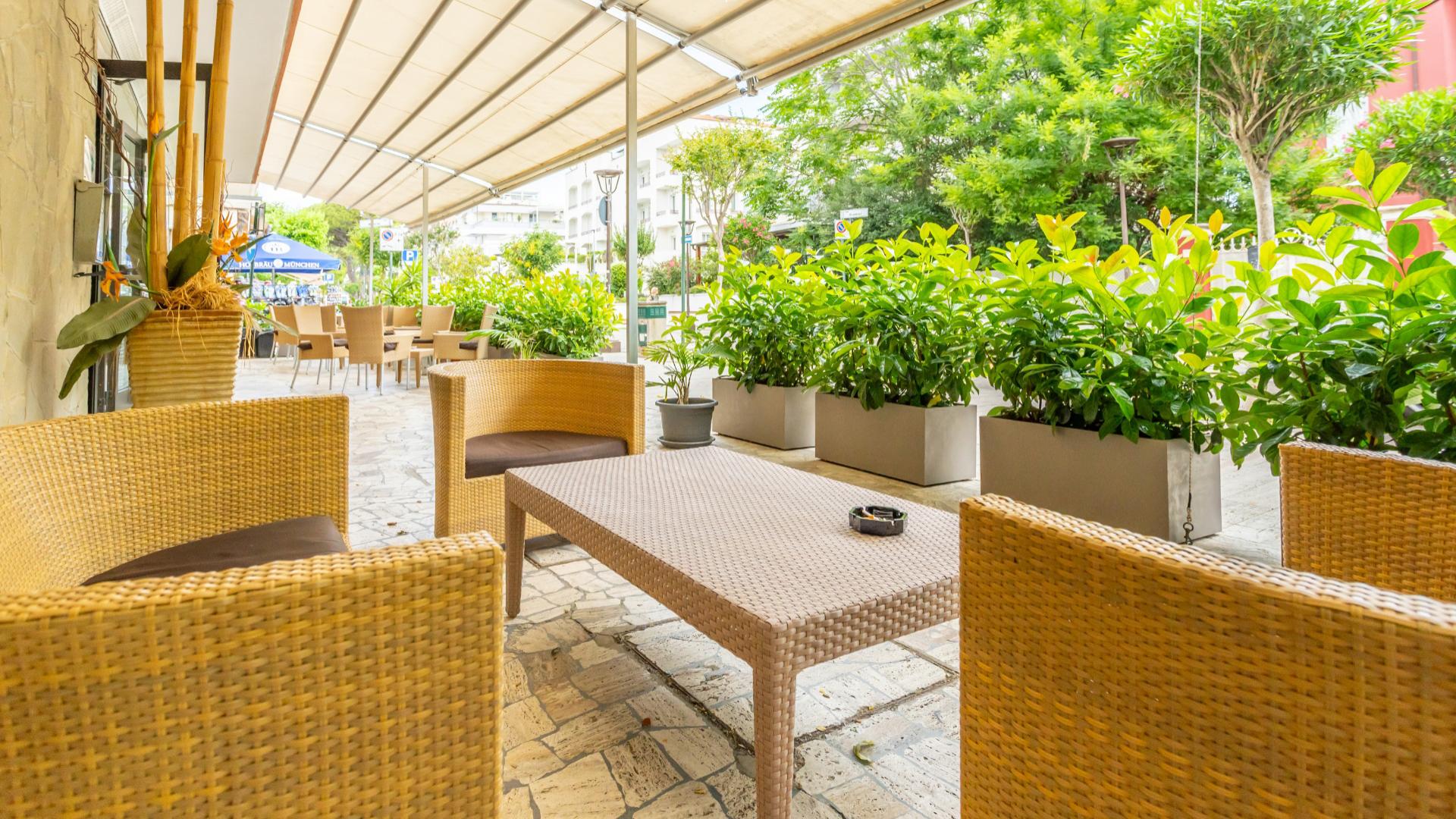 Terrazza con tavoli e sedie in vimini, circondata da piante verdi.