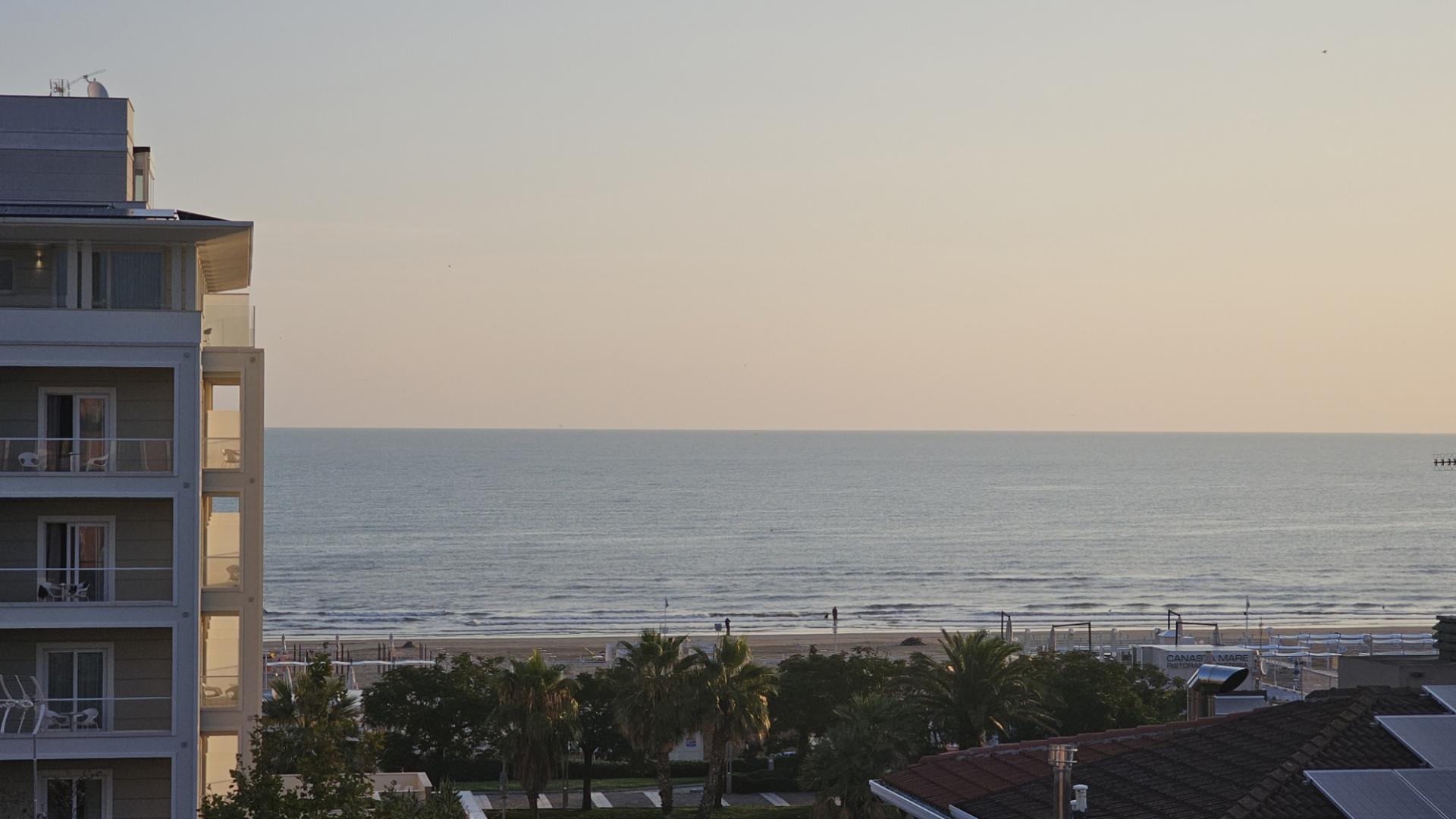 Vista sul mare al tramonto con un edificio sulla sinistra e palme in primo piano.