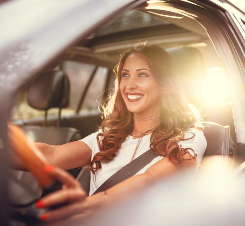 Donna sorridente guida un'auto al tramonto, luce solare illumina l'interno.