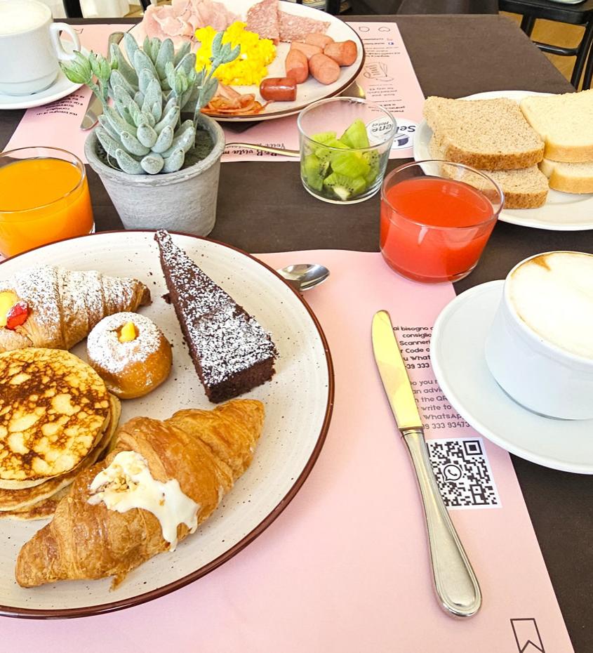Colazione ricca con dolci, frutta, succhi e cappuccino su tavola apparecchiata.